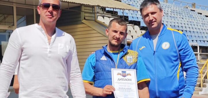 Троє людей стоять на стадіоні. У центрі — спортсмен тримає диплом. Двоє в спортивних костюмах із написом «Ukraine».