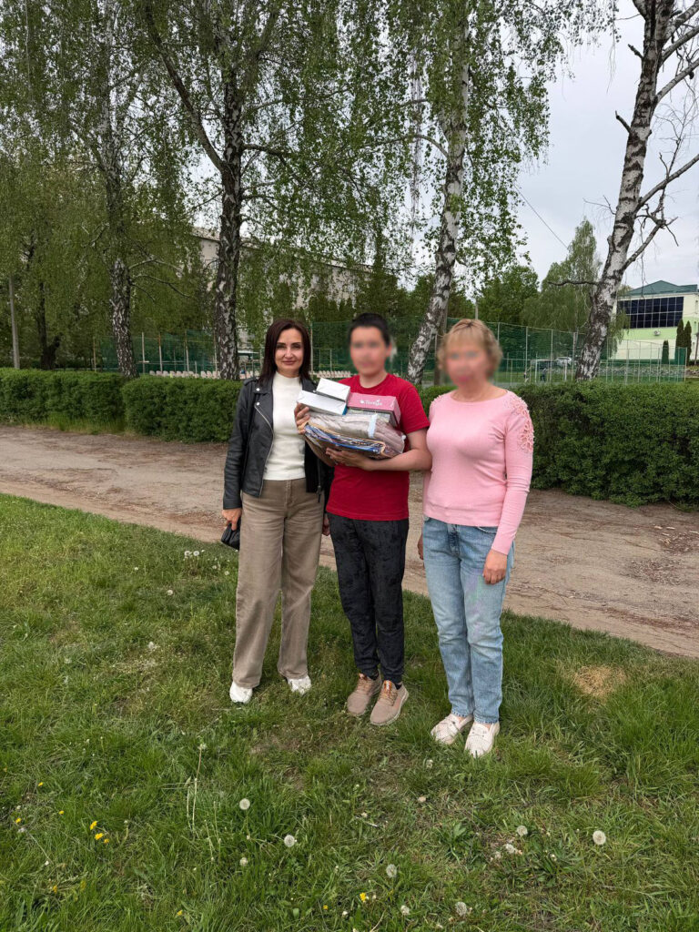 Троє людей стоять разом на траві. У центрі — людина з кількома предметами в руках. Позаду — дерева, будівля та огорожа.