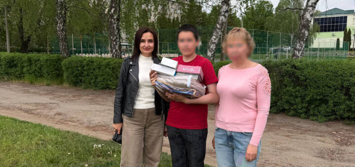 Троє людей стоять разом на траві. У центрі — людина з кількома предметами в руках. Позаду — дерева, будівля та огорожа.