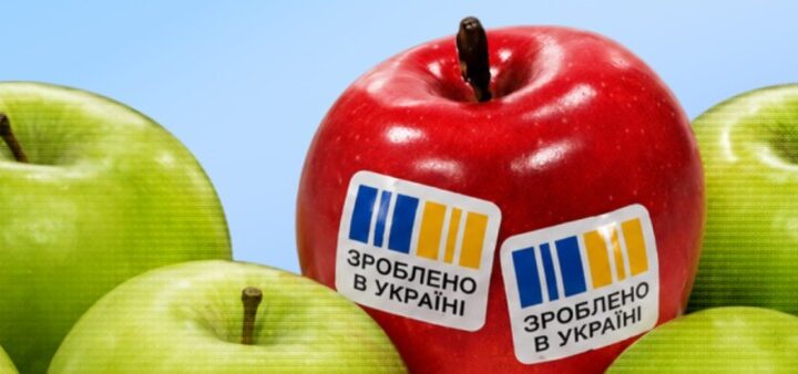 На блакитному фоні група яблук: більшість зелені, а в центрі червоне яблуко зі стікерами “Зроблено в Україні”