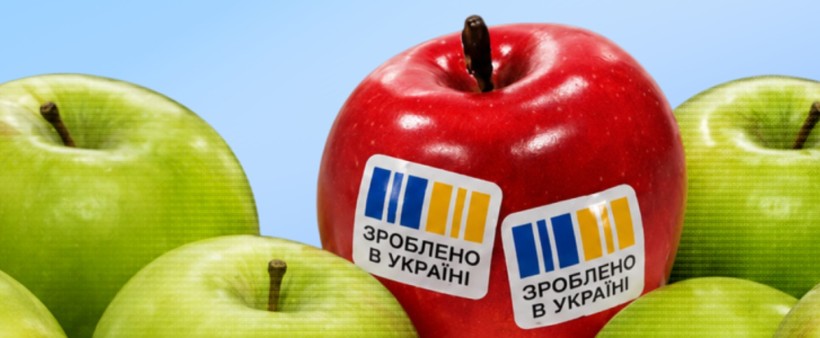 На блакитному фоні група яблук: більшість зелені, а в центрі червоне яблуко зі стікерами “Зроблено в Україні”