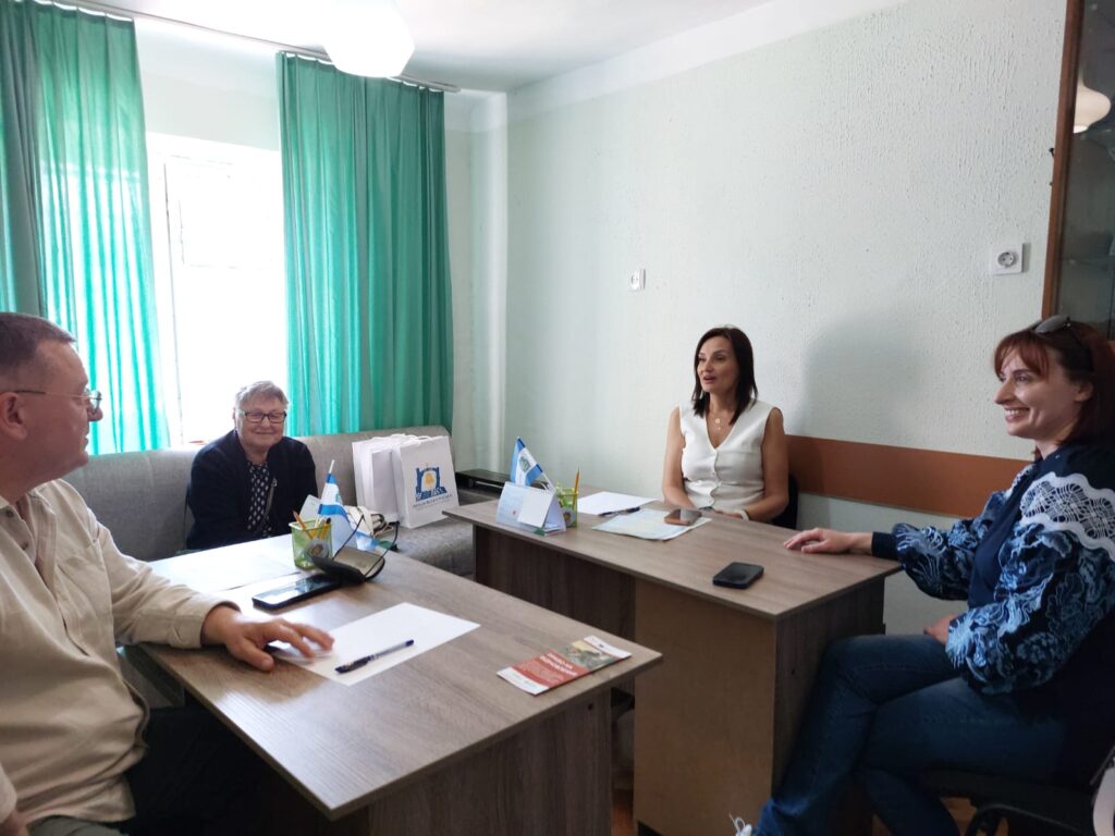 Четверо учасників сидять за двома дерев’яними столами в невеликій кімнаті. На столах — папери, телефони, маленькі прапорці та канцелярія. Позаду — подарункові пакети з синьо-жовтим логотипом.