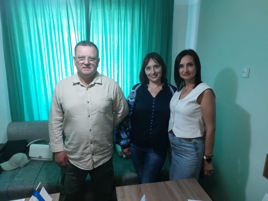 Троє учасників позують перед бірюзовою шторою. Ліворуч — чоловік у бежевій сорочці, посередині — жінка в темно-синій блузі, праворуч — жінка у світлому топі та джинсах.