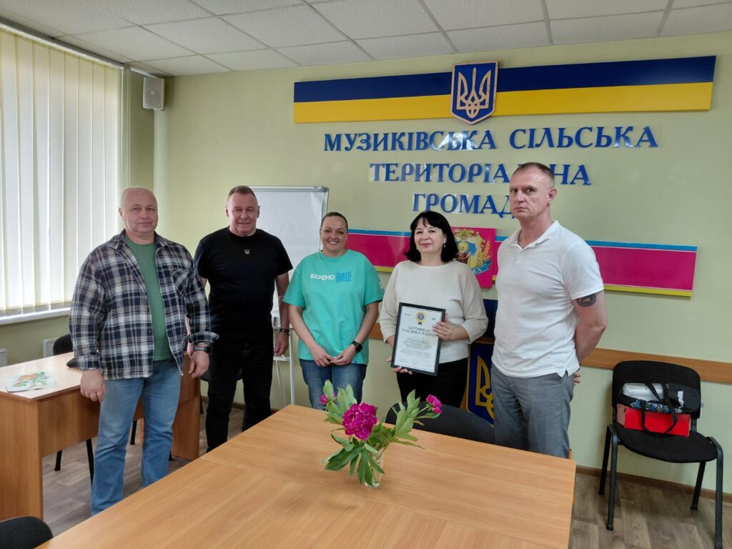П’ятеро людей стоять у кімнаті з зеленими стінами. Позаду — герб України та напис «Музиківська сільська територіальна громада». Один учасник тримає рамку з сертифікатом. На столі попереду — ваза з рожевими квітами.