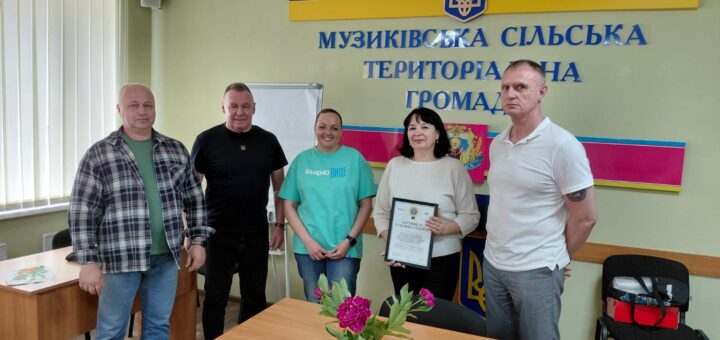 П’ятеро людей стоять у кімнаті з зеленими стінами. Позаду — герб України та напис «Музиківська сільська територіальна громада». Один учасник тримає рамку з сертифікатом. На столі попереду — ваза з рожевими квітами.