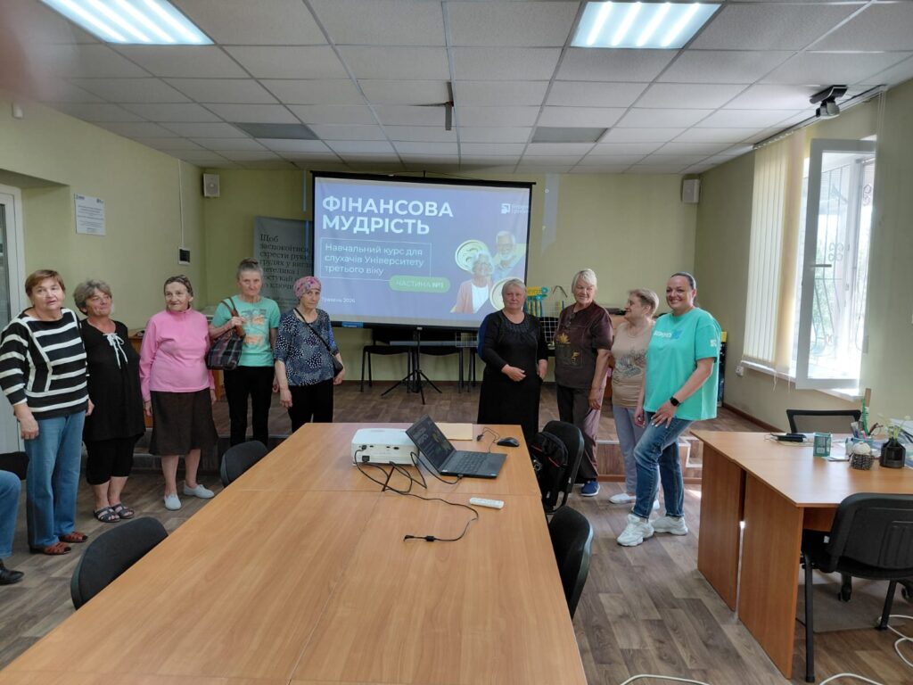 Група літніх людей стоїть у класі. На екрані проєктора — слайд із написом «ФІНАНСОВА МУДРІСТЬ. Навчальний курс для слухачів Університету третього віку. Частина №1»