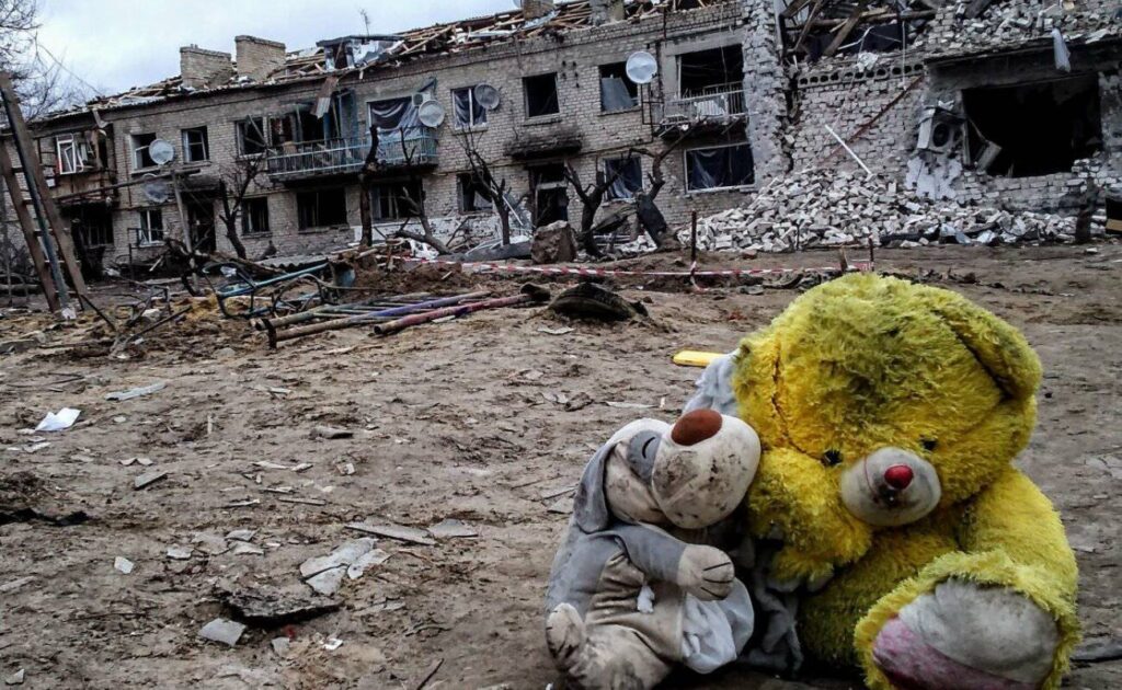 Дві м’які іграшки — жовтий ведмедик і менша сіра тваринка — сидять на землі перед зруйнованим будинком із вибитими вікнами та уламками