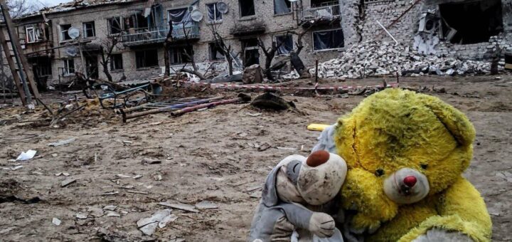 Дві м’які іграшки — жовтий ведмедик і менша сіра тваринка — сидять на землі перед зруйнованим будинком із вибитими вікнами та уламками