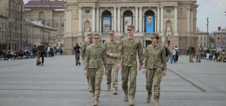 П’ятеро військових у камуфляжі йдуть площею перед величною будівлею з колонами та статуями; навколо цивільні люди відпочивають і пересуваються