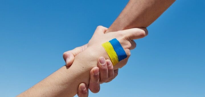 Дві руки міцно тримають одна одну на фоні ясного блакитного неба; на зап’ясті однієї руки намальований синьо‑жовтий прапор України