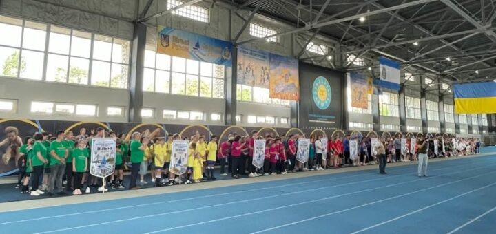 У просторому приміщенні з біговою доріжкою стоять групи людей у різнокольорових футболках — зелених, жовтих, червоних, білих. Вони вишикувані в ряди, тримають банери, серед них напис «Вільні»