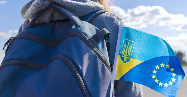 Людина у сірій худі з синім рюкзаком; на рюкзаку маленький прапор із поєднанням українських синьо‑жовтих смуг, тризуба та жовтих зірок ЄС; на тлі ясне небо з хмарами