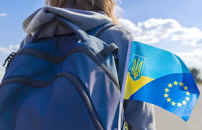 Людина у сірій худі з синім рюкзаком; на рюкзаку маленький прапор із поєднанням українських синьо‑жовтих смуг, тризуба та жовтих зірок ЄС; на тлі ясне небо з хмарами