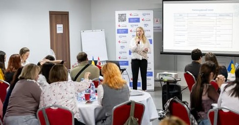 Конференц‑зал із людьми за столами на червоних стільцях; доповідач із мікрофоном стоїть біля екрана з таблицею “Technological areas”; позаду банер із логотипами та текстом