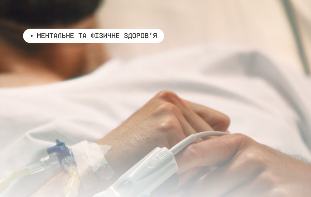 Рука людини з медичним сенсором на пальці та крапельницею у вені; пацієнт лежить у лікарняному ліжку в білому халаті, на тлі розмитий інтер’єр