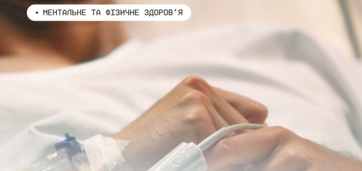 Рука людини з медичним сенсором на пальці та крапельницею у вені; пацієнт лежить у лікарняному ліжку в білому халаті, на тлі розмитий інтер’єр