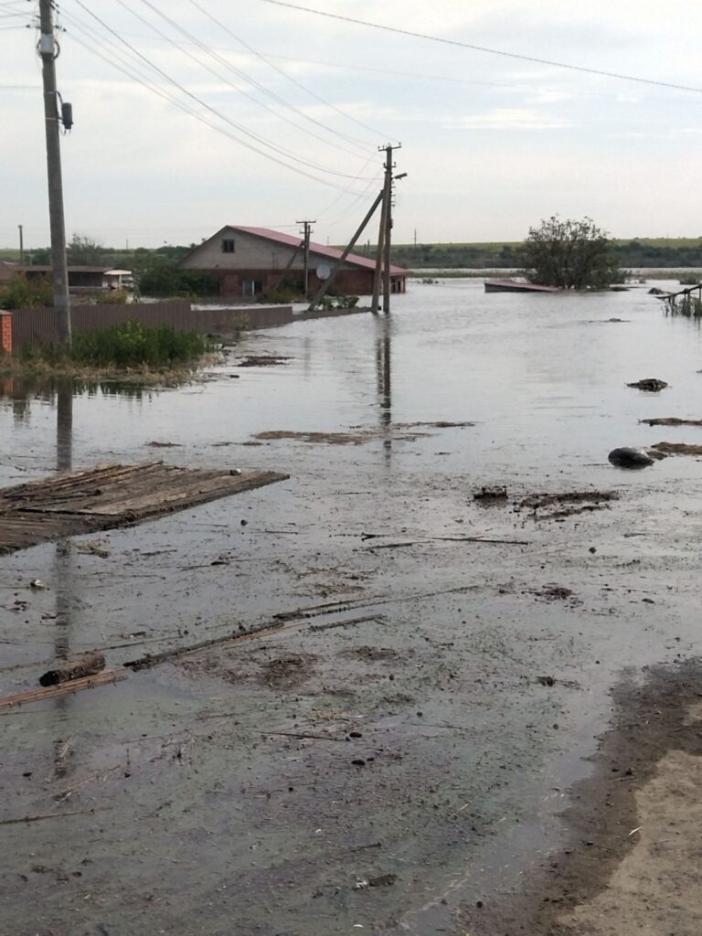 Затоплена місцевість із дорогою та електричними стовпами, частково зануреними у воду; на задньому плані будівлі з підтопленими стінами