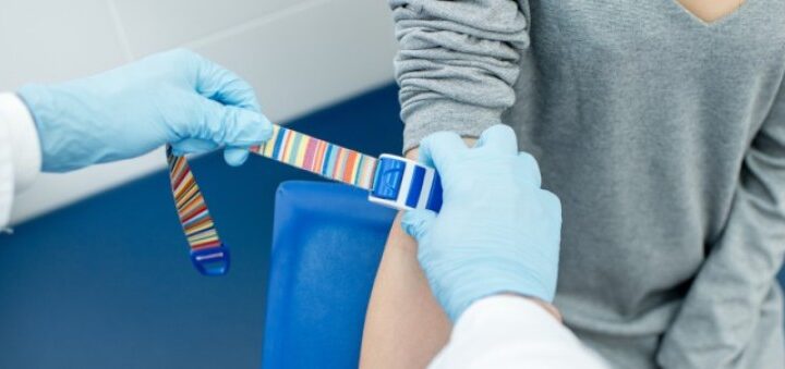 Медичний працівник у синіх рукавичках накладає різнокольоровий джгут на руку пацієнта у сірій кофті; підготовка до забору крові чи ін’єкції.