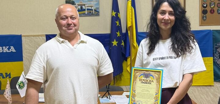 Михайло Жадан та БОРТОВИК Лариса стоять всвітлому кабінеті. Жінка тримає в руках подяку, позаду них прапори України та Євросоюзу