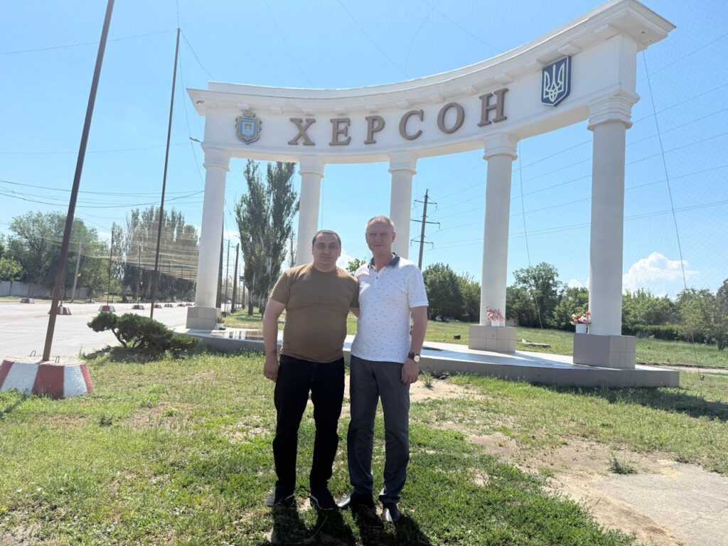 Двоє людей стоять перед білою архітектурною спорудою з високими колонами та написом «ХЕРСОН». Ліворуч — герб, праворуч — тризуб. Позаду — дерева, електролінії та ясне синє небо. 