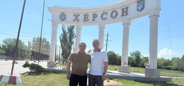 Двоє людей стоять перед білою архітектурною спорудою з високими колонами та написом «ХЕРСОН». Ліворуч — герб, праворуч — тризуб. Позаду — дерева, електролінії та ясне синє небо.