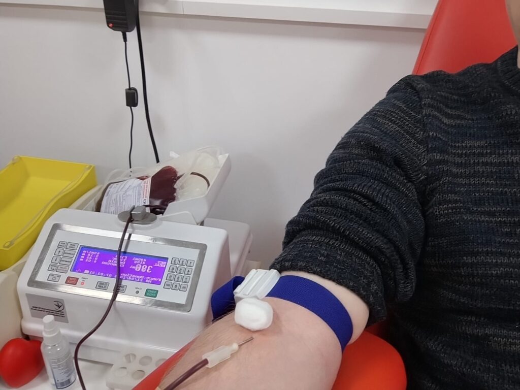 Рука людини на червоному кріслі, накладений синій джгут, у вену введена голка з трубкою, що веде до пакета для збору крові. Поруч — медичний апарат із цифровим дисплеєм, контейнер та дезінфектор