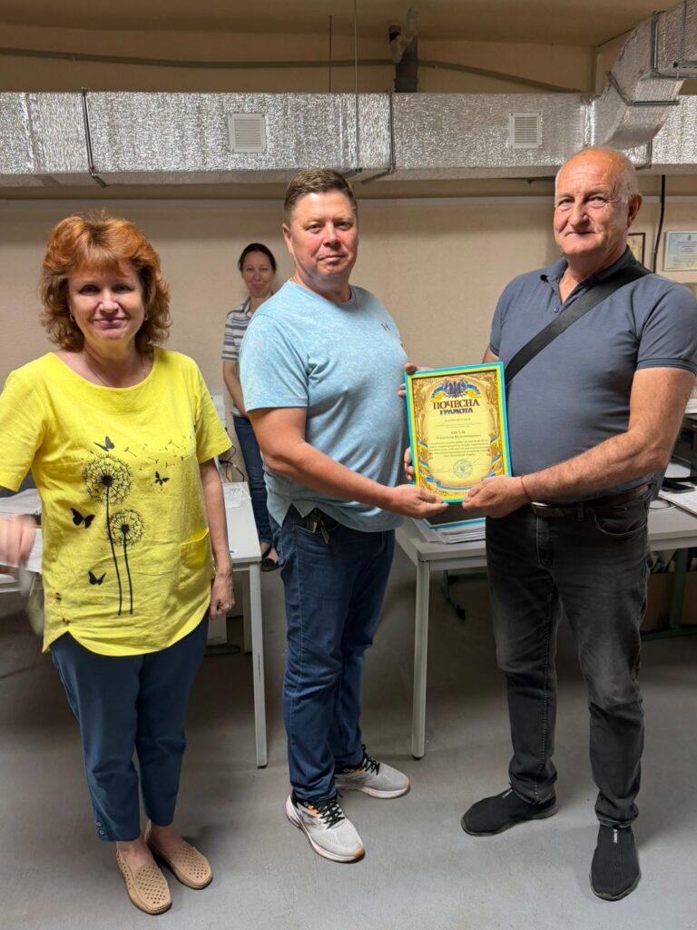Четверо людей стоять у приміщенні з офісними столами. Двоє тримають кольоровий сертифікат із написом «Почесна грамота».
