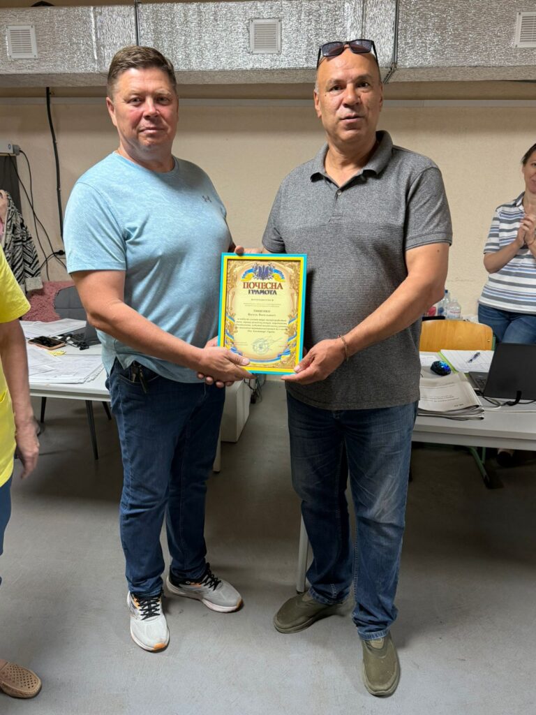 Двоє людей тримають сертифікат із синьо-жовтою рамкою та тризубом угорі, текст — «Подяка». Позаду — столи з ноутбуками, паперами та офісним обладнанням. 