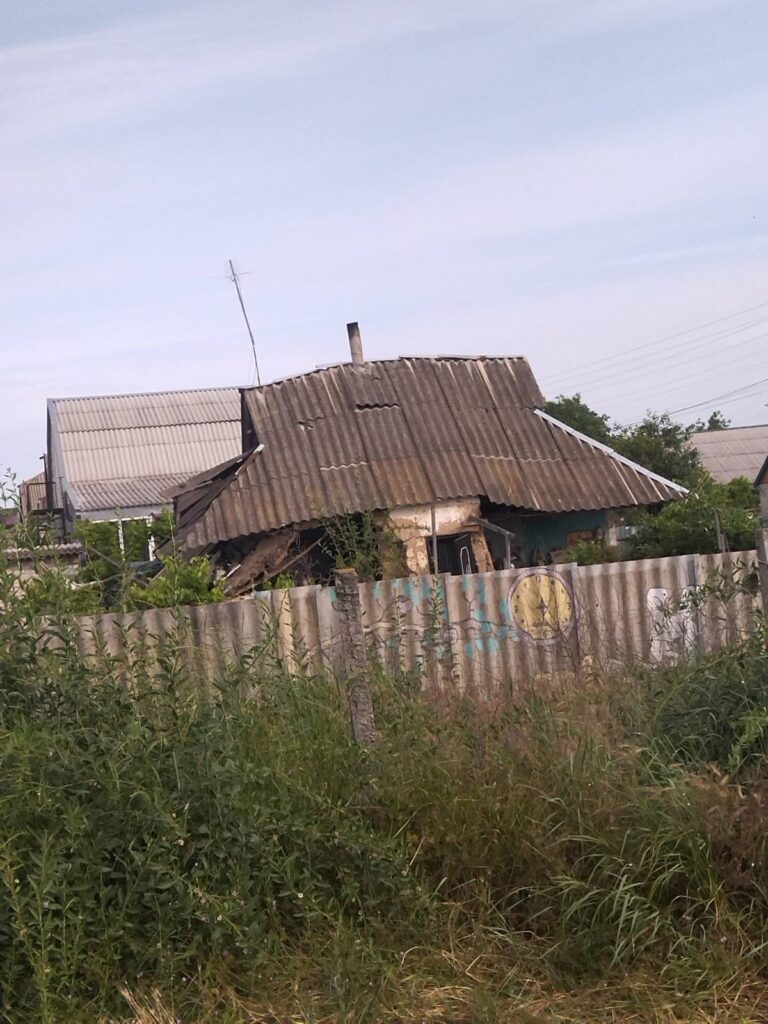 Старий занедбаний будинок із частково обваленим дахом та іржавим металевим парканом із бляклими написами; довкола заросла рослинність