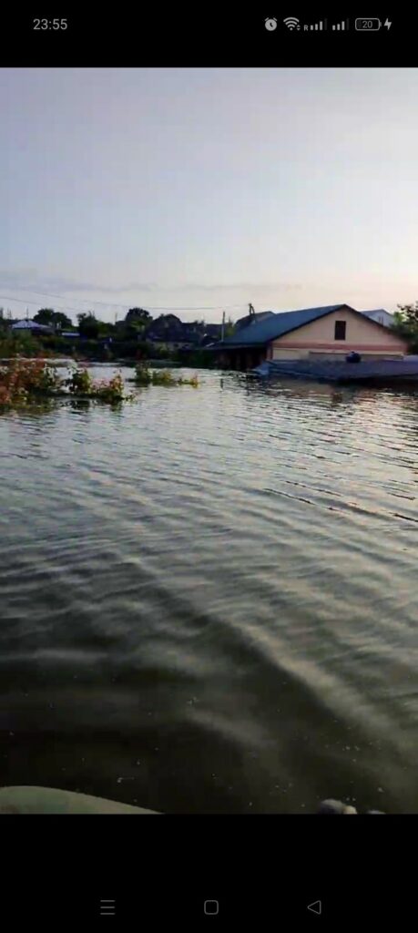 «Затоплений будинок із блакитним дахом, вода сягає стін; навколо видно інші споруди та рослинність, частково занурені у воду