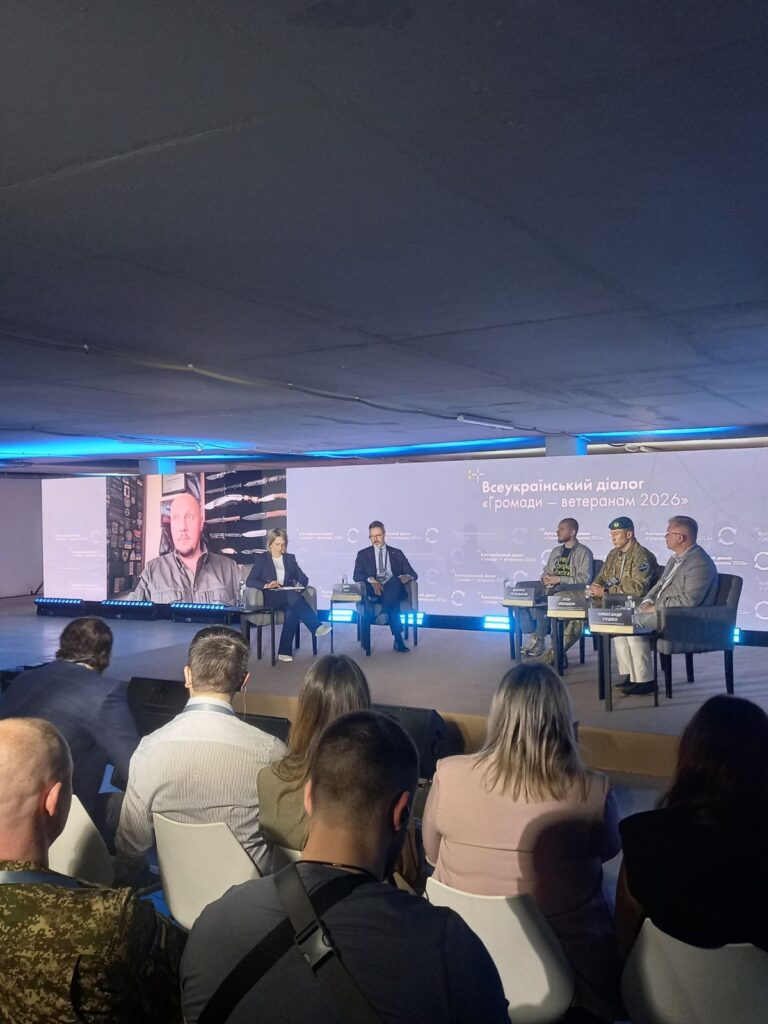 Знімок великої зали з аудиторією, яка сидить перед сценою. На сцені — п’ятеро учасників, серед них двоє у військовій формі, ще один виступає дистанційно на великому екрані. Позаду — синій фон із написом «Всеукраїнський діалог “Громади – ветеранам 2026”».