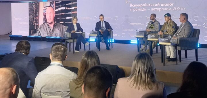 Знімок великої зали з аудиторією, яка сидить перед сценою. На сцені — п’ятеро учасників, серед них двоє у військовій формі, ще один виступає дистанційно на великому екрані. Позаду — синій фон із написом «Всеукраїнський діалог “Громади – ветеранам 2026”».