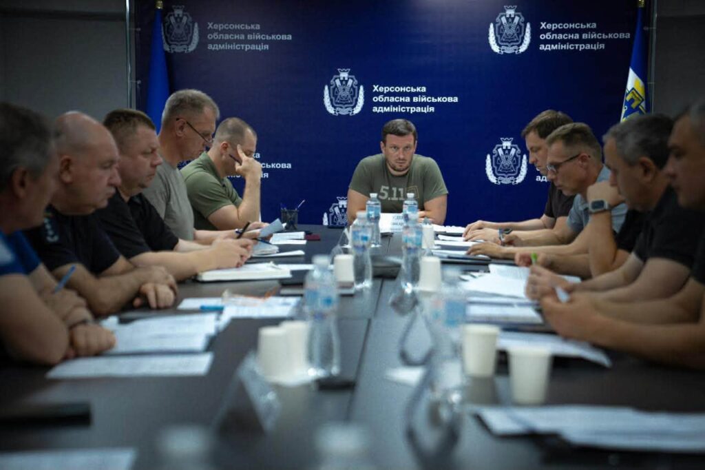 Велика нарада за конференц-столом, учасники з блокнотами та документами. На задньому плані — синій банер із написом «Херсонська обласна військова адміністрація» та офіційним гербом.
