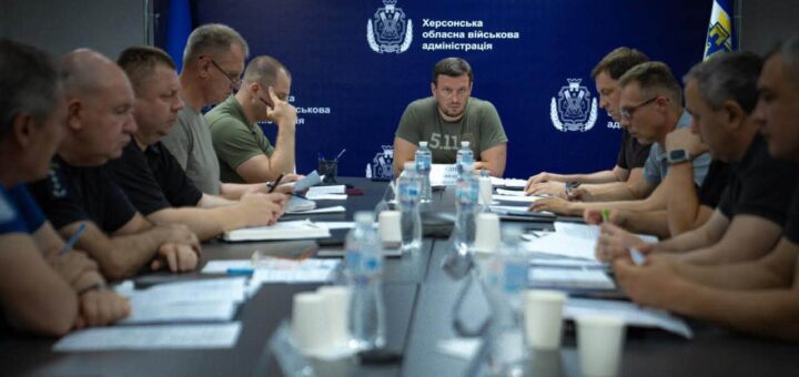 Велика нарада за конференц-столом, учасники з блокнотами та документами. На задньому плані — синій банер із написом «Херсонська обласна військова адміністрація» та офіційним гербом.