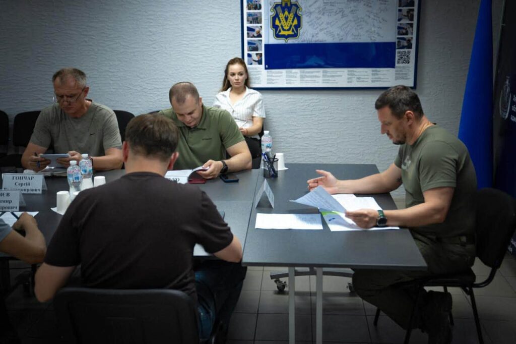 На засіданні за прямокутним столом сидять учасники засідання, перед ними таблички з іменами. На стіні — великий синьо-білий плакат із жовто-блакитним символом, підписами та фото.
