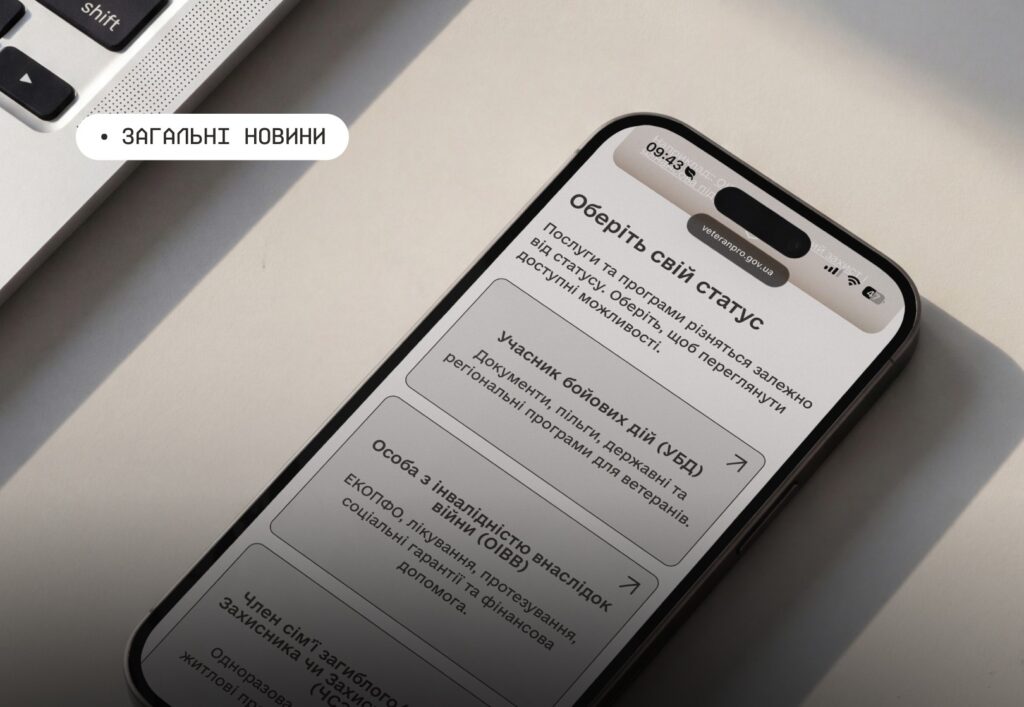Смартфон на світлій поверхні поруч із клавіатурою ноутбука; на екрані сторінка “Оберіть свій статус” із трьома категоріями підтримки для ветеранів та їхніх родин