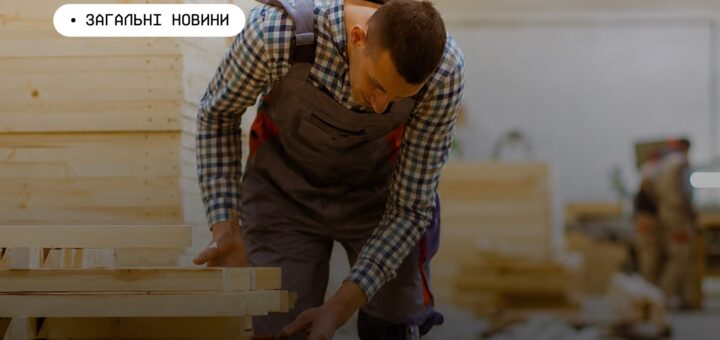 юдина у майстерні в картатій сорочці та комбінезоні нахиляється над стопкою дерев’яних дощок; позаду видно інші стоси деревини та ще одного працівника; текст “Загальні новини” українською