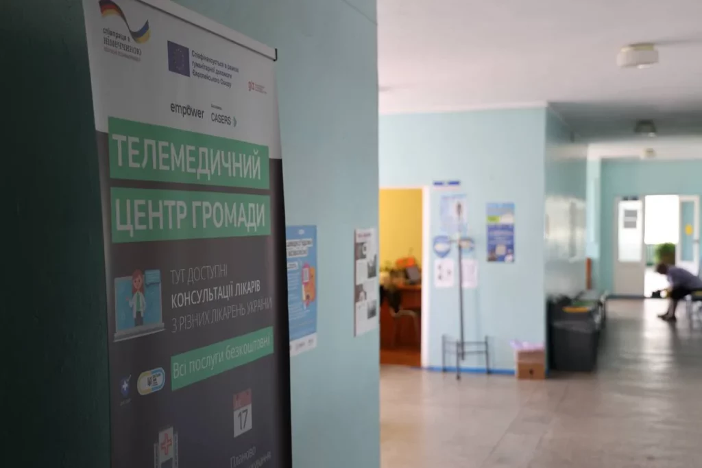 Інтер’єр медичного закладу з блакитними стінами та коридором, що веде до кількох кабінетів. Ліворуч — вертикальний банер із написом «Телемедичний центр громади» та логотипами ЄС, GIZ, empower, CASERS. Текст на банері повідомляє, що консультації лікарів із різних лікарень України доступні безкоштовно. У глибині коридору — плакати на стінах, відкрита двері з робочим столом та людина, яка сидить на лавці.