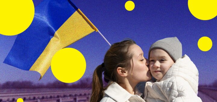 Жінка цілує дитину в щоку, тримаючи її на руках. Обоє в світлих пальтах, дитина в сірій в’язаній шапці. На тлі — синьо-жовтий прапор України, що майорить на чистому небі, та розмитий пейзаж із мостом чи перилам