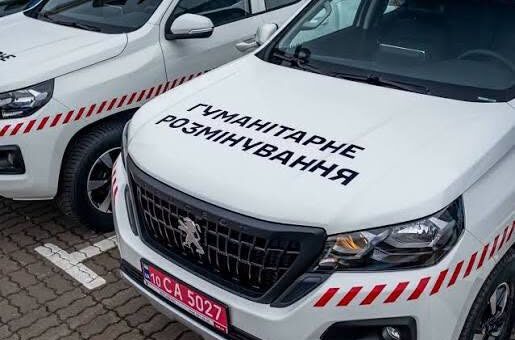 Дві білі спеціалізовані машини з червоно‑білими діагональними смугами на капоті та бортах; на передньому автомобілі напис українською “Гуманітарне розмінування”; червоний номерний знак із білими літерами; транспортні засоби припарковані на асфальтованій поверхні