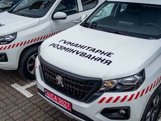 Дві білі спеціалізовані машини з червоно‑білими діагональними смугами на капоті та бортах; на передньому автомобілі напис українською “Гуманітарне розмінування”; червоний номерний знак із білими літерами; транспортні засоби припарковані на асфальтованій поверхні