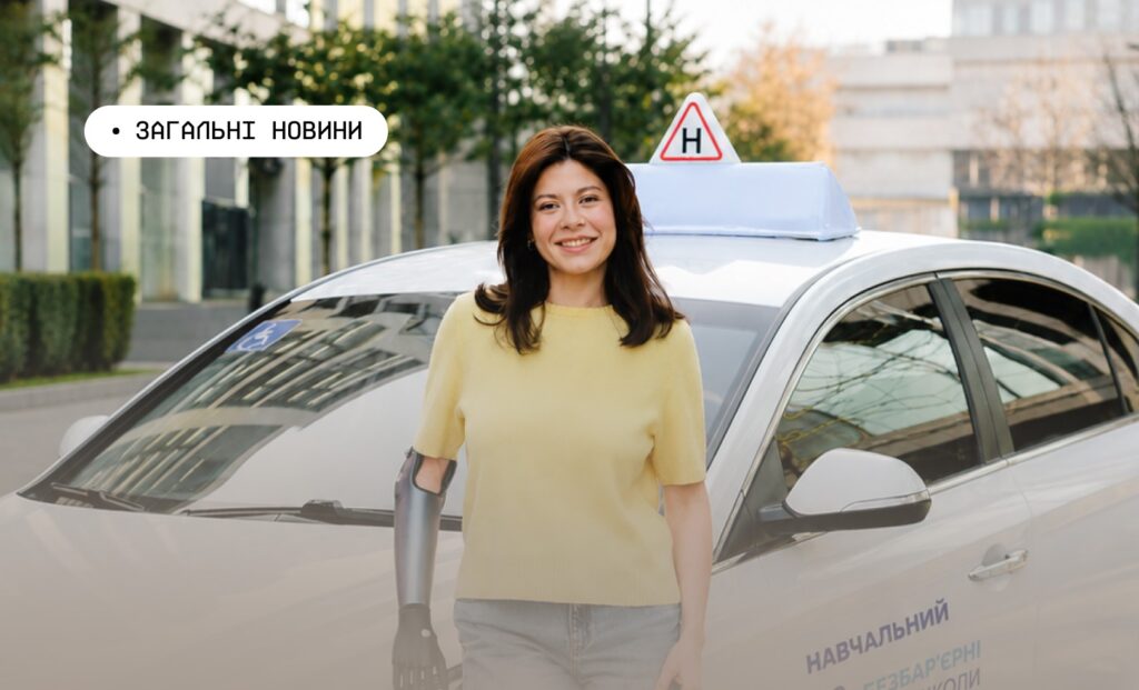 Людина з протезом руки стоїть перед білим автомобілем із позначкою “H” на даху та символом інвалідності на лобовому склі; на боці машини напис українською “Навчальний”; на фоні сучасні будівлі, зелень і вимощена площа