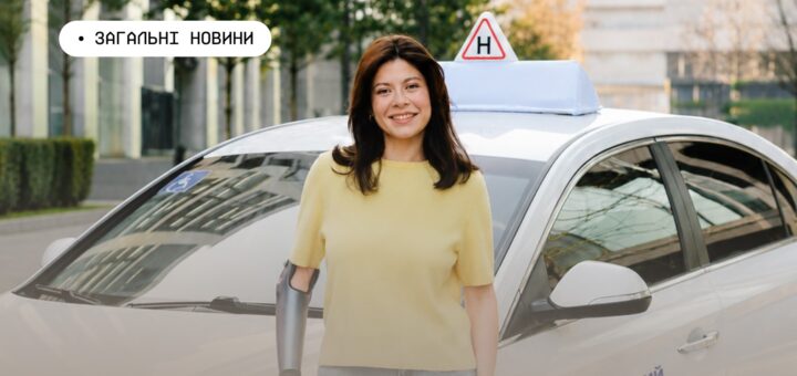 Людина з протезом руки стоїть перед білим автомобілем із позначкою “H” на даху та символом інвалідності на лобовому склі; на боці машини напис українською “Навчальний”; на фоні сучасні будівлі, зелень і вимощена площа