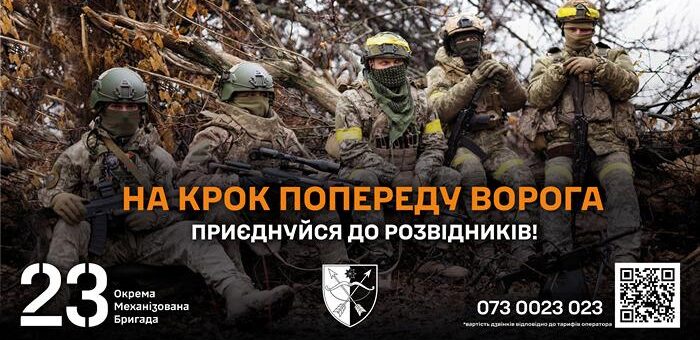 П’ятеро військових у камуфляжі та тактичному спорядженні сидять або присіли серед сухих гілок у лісистій місцевості. Усі зі зброєю, у шоломах та захисних окулярах. Текст: «На крок попереду ворога. Приєднуйся до розвідників! 23 Окрема Механізована Бригада. 073 0023 023». Праворуч — чорнобілий щит‑емблема та QR‑код.