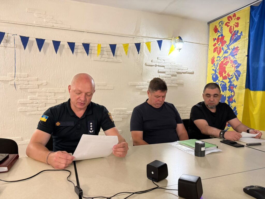 За світлис столом 3 учасника нараби у темному одязі, один з них читає доповідь, позаду - світла кам'яна стіна підвального приміщення