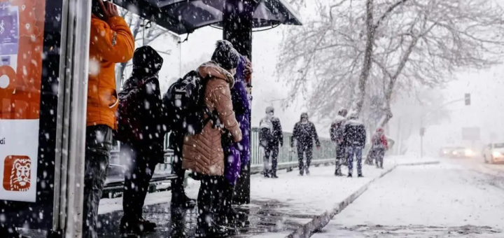 Зимова міська сцена: люди в теплих пальтах і шапках стоять на зупинці автобуса та йдуть засніженим тротуаром; падає сніг, дерева без листя, автомобілі з увімкненими фарами на тлі; атмосфера холодна й буденна.