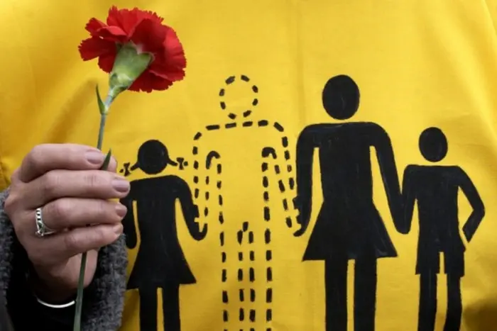 На жовтій футболці зображена чорна графіка сім’ї з чотирьох осіб, одна фігура позначена пунктиром — символ відсутності. У руці людина тримає червону гвоздику.