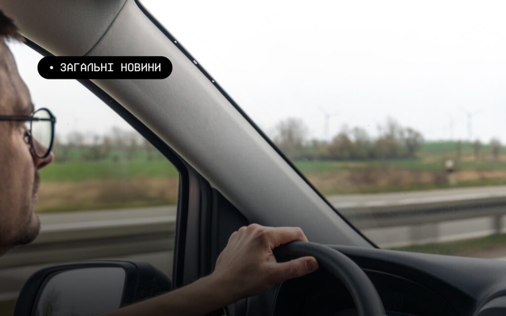 Інформаційне фото: людина веде автомобіль, видно руку на кермі, дзеркало та частину панелі; за вікном — трасове огородження, зелені поля й вітрові турбіни на горизонті під похмурим небом; текст — “ЗАГАЛЬНІ НОВИНИ”