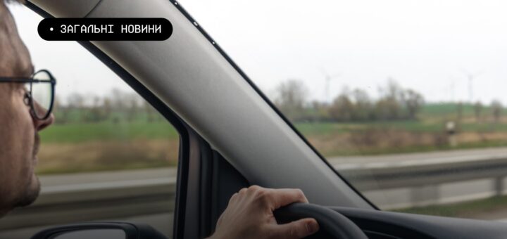 Інформаційне фото: людина веде автомобіль, видно руку на кермі, дзеркало та частину панелі; за вікном — трасове огородження, зелені поля й вітрові турбіни на горизонті під похмурим небом; текст — “ЗАГАЛЬНІ НОВИНИ”