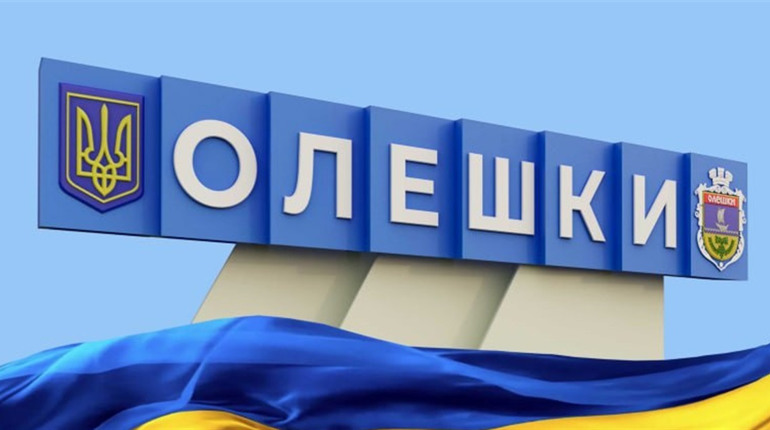 Інформаційний постер українською мовою: великий синьо‑білий знак із написом “ОЛЕШКИ”; ліворуч тризуб — державний герб України; праворуч герб міста; на передньому плані майорить синьо‑жовтий прапор; фон — ясне блакитне небо